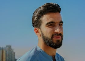 A close-up outdoor portrait of a young man with facial hair, exuding confidence under a clear sky.